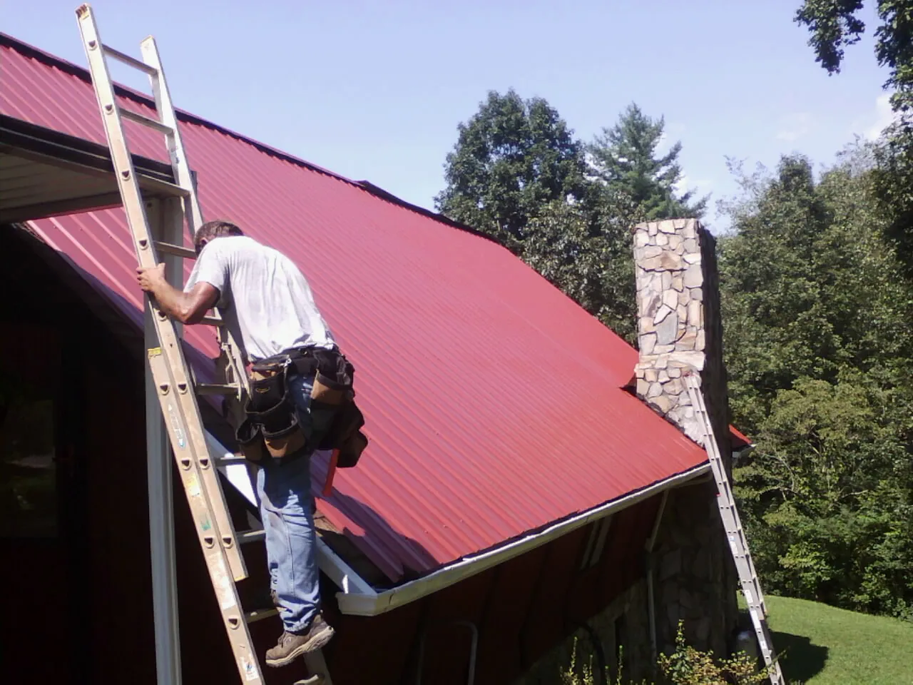 Roofer on a ladder beside a red metal roof and stone chimney — Soffit & Fascia Repair by Roofing Brothers in Rossburg